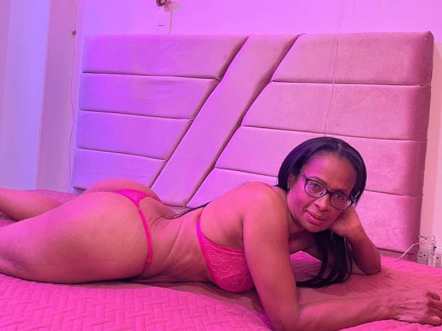 AfricaDLuxe está acostada en una cama rosa, vistiendo lencería de encaje rosa, con las nalgas levantadas, mirando a la cámara, y lleva gafas, con iluminación rosa..