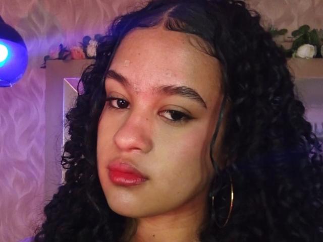VelaskaJones has curly black hair, medium brown skin, and red lipstick with small water droplets on her forehead. She's wearing large gold hoop earrings and has subtle eye makeup.