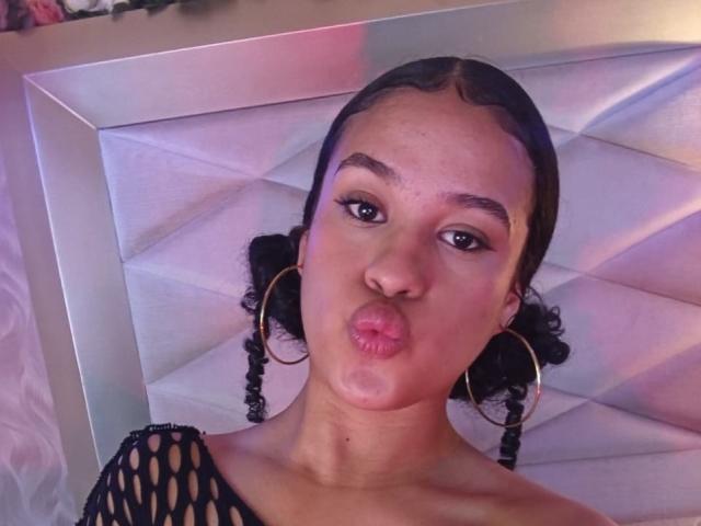 VelaskaJones, avec ses lèvres roses et gonflées, son haut en résille noire, ses boucles d'oreilles dorées et ses cheveux tressés, faisant une moue coquine, allongée sur une tête de lit blanche et matelassée..