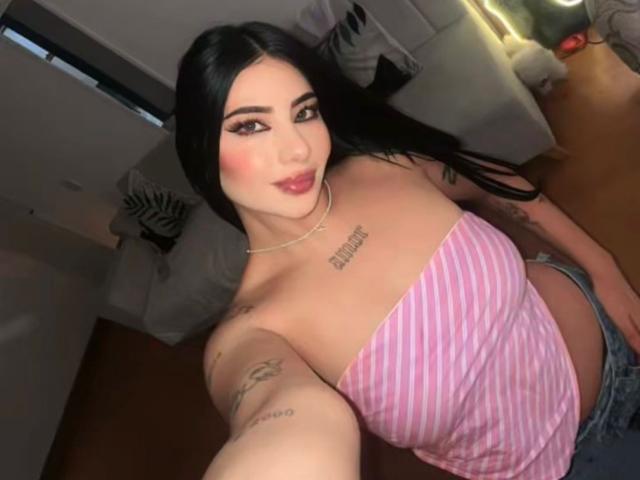 NatallieBlue with black hair, pink striped top, tattoos, and red lipstick, showing her tits, wearing denim jeans, taking a selfie in a dimly lit room.