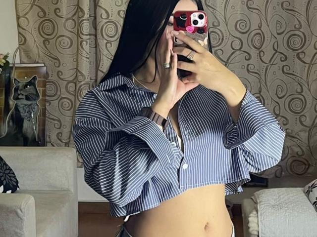 NatallieBlue mit langen, schwarzen Haaren, trägt ein blau gestreiftes Crop-Top, hält ein rotes Telefon, zeigt ihren Bauch und ihre kleinen Brüste, wobei ihre Hand ihren Mund bedeckt..