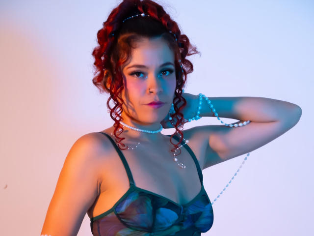 Arielle Brooks, avec ses cheveux roux bouclés, portant un soutien-gorge en mesh vert, un collier de perles bleues et un bracelet de perles bleues, regardant l'appareil photo avec une main derrière la tête..