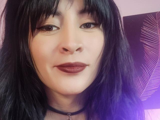 IssisDiosa has pale skin, black straight hair with bangs, dark eyeliner, and red lipstick. She wears a black choker and looks directly at the camera with a slight smile.