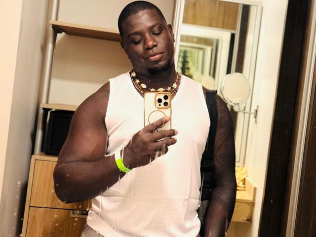 Darren Stone, peau foncée, bras musclés, cheveux courts, chemise blanche sans manches, collier en or, bracelet vert, tenant un téléphone, prenant un selfie devant un miroir, souriant, reflet dans le miroir, décor d'une chambre..