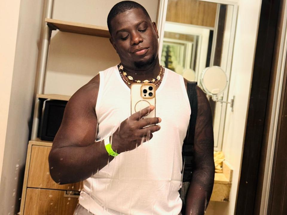 DarrenStone with dark skin, muscular arms, short haircut, white sleeveless shirt, gold necklace, green wristband, holding phone, taking mirror selfie, smiling, mirror reflection, bedroom setting.