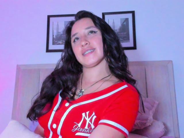 JessyBarnnett with long black hair, wearing a red NY Jets shirt, smiling, necklace with a heart pendant, sitting on a beige bed, blue and pink lighting.