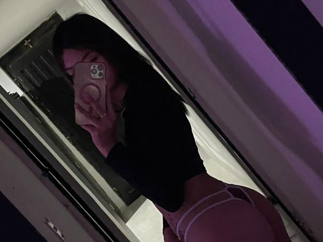 ScarlettPreatty with black hair, wearing a black top and pink thong, showing her round ass in a mirror selfie, holding a phone with a pink case.