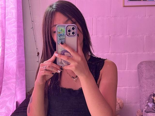 ScarlettPreatty taking a selfie, her phone covering her face, wearing a black lace top, pinkish lighting, visible tattoo on her right forearm, pink wall, and a purple curtain.