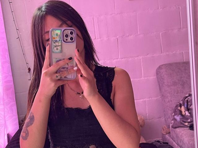 ScarlettPreatty with long brown hair, black lace sleeveless top, holding phone covering face, pink light, black nails, tattoo on right forearm, necklace, purple-gray chair, mirror selfie.