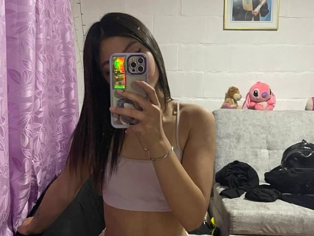 ScarlettPreatty taking a mirror selfie, wearing a pink sports bra, showing her small tits, holding a phone with a colorful case, in a room with purple curtains.