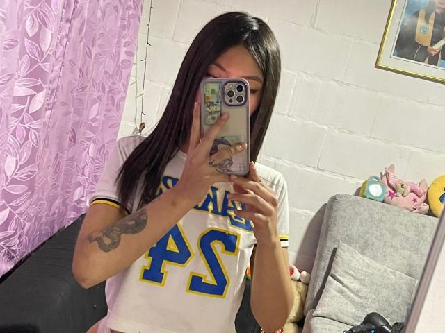 ScarlettPreatty with long black hair, wearing a white "PS" sports shirt, taking a mirror selfie with her phone, showing a tattoo on her left arm, in a cozy room.