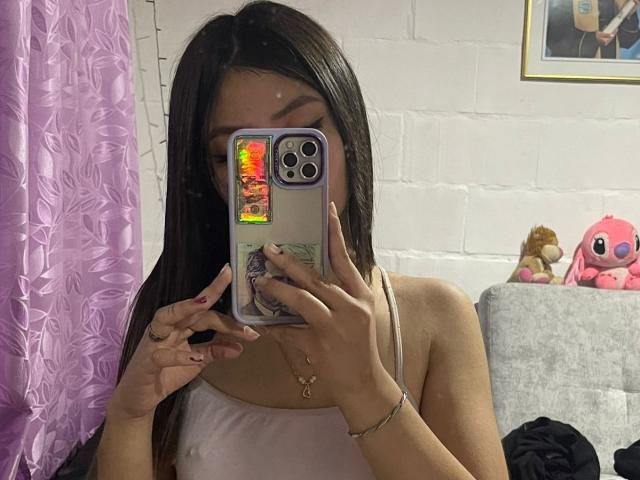 ScarlettPreatty with long black hair, taking a mirror selfie with her iPhone, showing her tits in a white tank top, pink nail polish, and a bracelet.