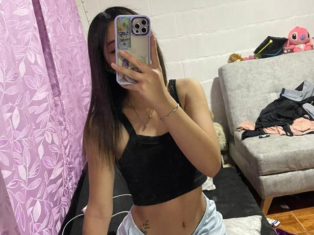 ScarlettPreatty with long brown hair, black crop top, light blue pajama bottoms, holding phone, showing midriff tattoo, pink curtain, gray sofa with clothes, and plush toys in background.