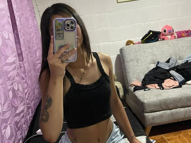 ScarlettPreatty with long brown hair, black tank top, and blue pants, showing her flat stomach and tattooed arm, taking a mirror selfie with her phone. Boobs are small.