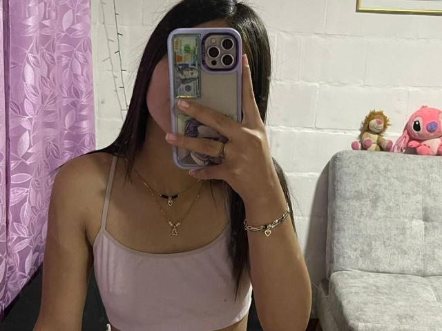 ScarlettPreatty with long black hair, pink tank top, gold necklaces, holding phone covering face, pink and purple room, grey couch with stuffed toys, string lights. Boobs visible.