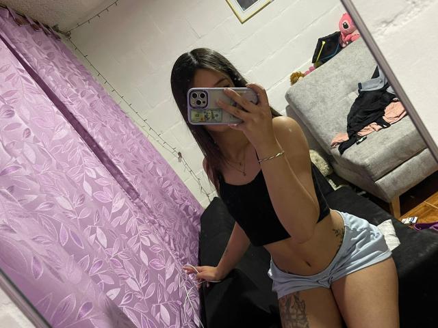 ScarlettPreatty with long brown hair, wearing a black crop top and light blue shorts, taking a mirror selfie, showing her flat stomach and tattooed thigh.