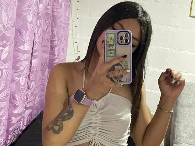 ScarlettPreatty with long black hair, wearing a white strapless top, holding a phone covering her face, showing a tattoo on her right arm, and a pink watch.