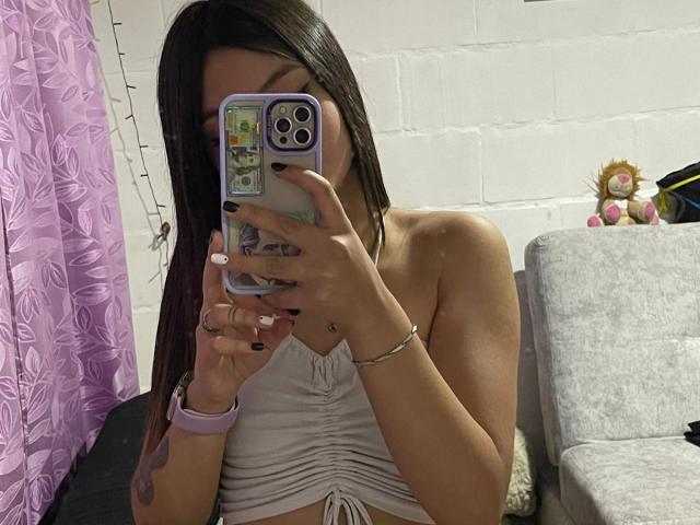 ScarlettPreatty with long brown hair, holding a phone covering her face, wearing a white tied-up top, showing her small tits, purple phone case, white bracelet, and pink wristwatch.