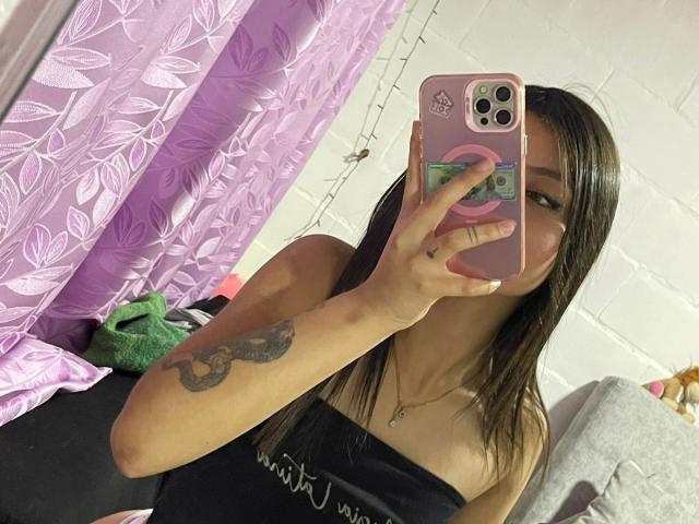 ScarlettPreatty with straight brown hair, black strapless top, holding pink phone with flower case, showing tattoo on right arm, taking mirror selfie, showing small tits.