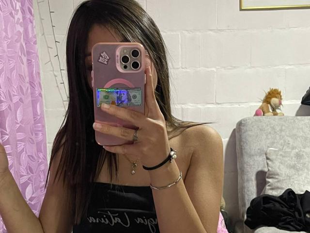 ScarlettPreatty with long brown hair, taking a mirror selfie with her pink phone, showing her bare shoulders, black top, and a ring on her right hand.