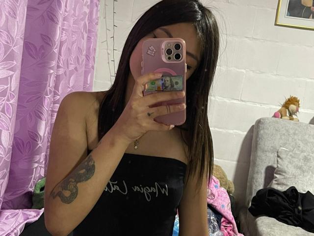 ScarlettPreatty with long brown hair, black strapless top, holding pink phone, showing tattoo on left arm, taking mirror selfie, pink curtain, white brick wall, stuffed toy on gray couch.