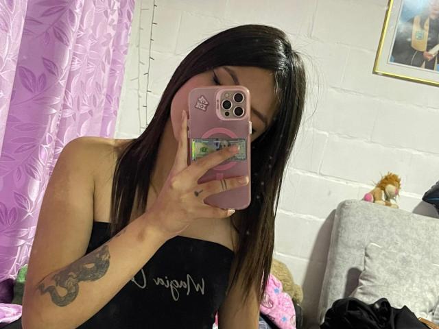 ScarlettPreatty with straight black hair, holding a pink phone covering her face, wearing a black strapless top, showing a tattoo on her right arm, and pink nails.