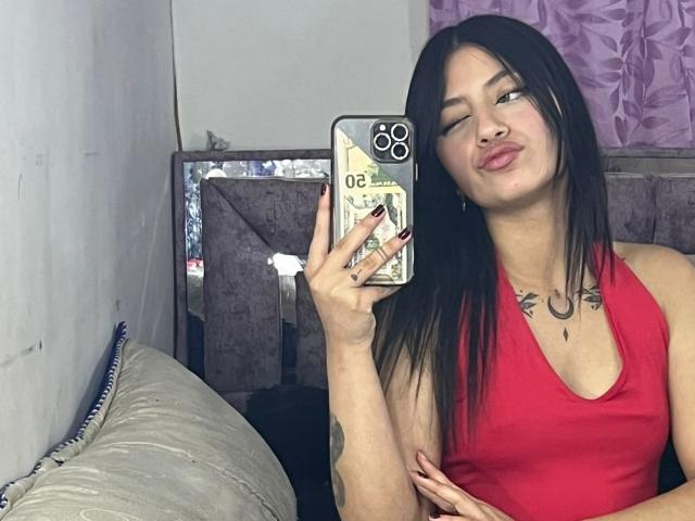 ScarlettPreatty with long black hair, red tank top, and tattoos, holding phone, puckering lips, looking at reflection, showing her tits, in mirror selfie with purple curtain.