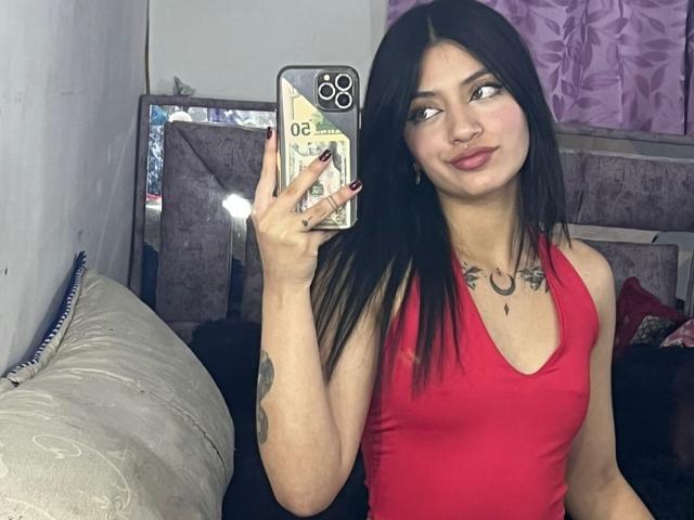 ScarlettPreatty with black hair, red tank top, and tattoos, taking a mirror selfie with a confident smile, showing her tits in the reflection.