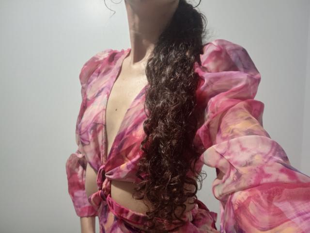 AngelRosa69 with curly brown hair, pink and purple tie-dye top, tied below her small tits, exposing her chest, standing against a plain white wall.