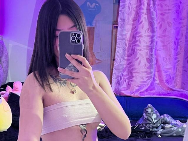 ScarlettPreatty taking a mirror selfie, showing off her bare tits wrapped in a white tube top, holding a phone, with purple lighting, and a tattoo on her chest.