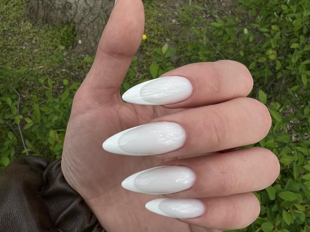 Adelisha's hand with super long, sharp, white acrylic nails, pointing up, showing off her shiny, glossy, and perfectly shaped nails against a green, leafy background.