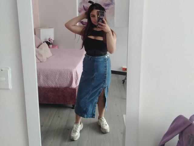 MelaniBecker in a mirror selfie, wearing a black cut-out top, blue high-waisted skirt with a thigh slit, white sneakers, and a purple hair clip, in a pink bedroom.