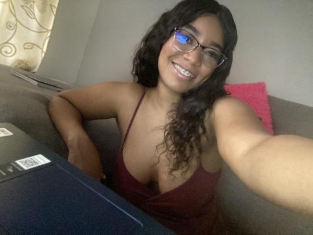 AvaJones69 with curly black hair, glasses, and a deep red tank top showing her tits, smiling at a camera while sitting on a gray couch. Laptop in front.