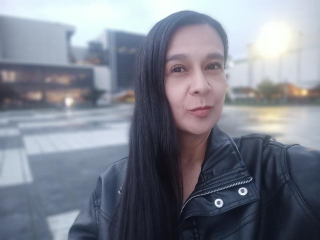 AileenRamirezW with long black hair, wearing a black leather jacket, has a subtle smile, and is standing outdoors in a blurry background. She's looking directly at the camera.