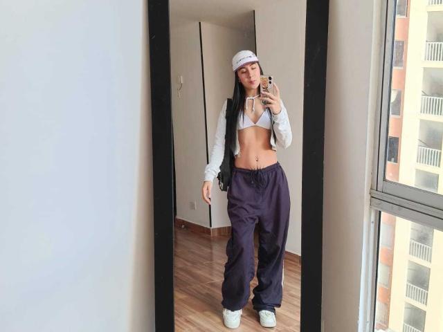 ChloeXk, con lunghi capelli neri, indossa una canotta bianca, pantaloni viola, scarpe da ginnastica bianche e un berretto bianco, e sta scattando un selfie allo specchio in una stanza con pavimenti in legno..