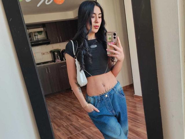 ChloeXk, con cabello largo y negro, un top corto negro, jeans azules, un bolso blanco y un teléfono, mostrando su abdomen descubierto y un piercing en el ombligo, de pie frente a un espejo..