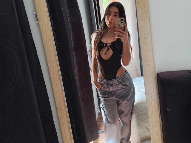 ChloeXk with long brown hair, black cutout top showing tits, and gray pants, taking a mirror selfie, hand in pocket, holding phone with pink case.