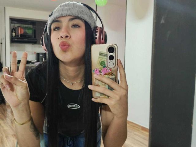 ChloeXk with long black hair, wearing a gray beanie and pink headphones, pouting her lips and making a peace sign with her right hand, holding a phone with stickers.