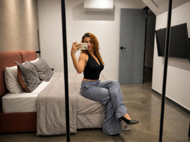 AriannaBlake with curly brown hair, black tank top, and blue high-waisted pants, sits on a bed, taking a mirror selfie, showing her ass in black heels.