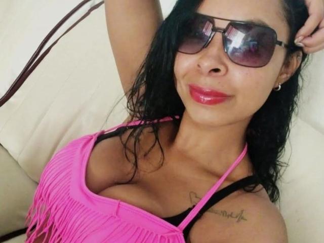Clair Bennet, com cabelo preto molhado, vestindo uma blusa rosa com franjas, alças pretas, seios grandes, batom rosa, óculos de sol escuros, sorrindo, braço direito levantado, tatuagem no ombro direito..