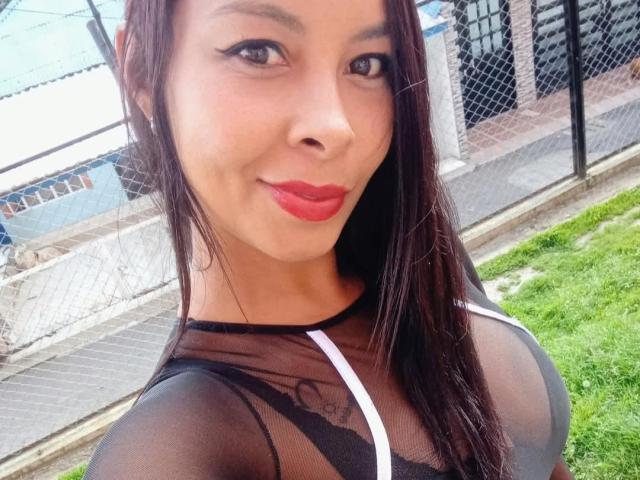 Clair Bennet, con lunghi capelli castani, rossetto rosso e una top nera trasparente che mette in mostra il suo seno prosperoso. Sorride, è in piedi all'aperto, con l'erba verde e una recinzione a catena sullo sfondo..