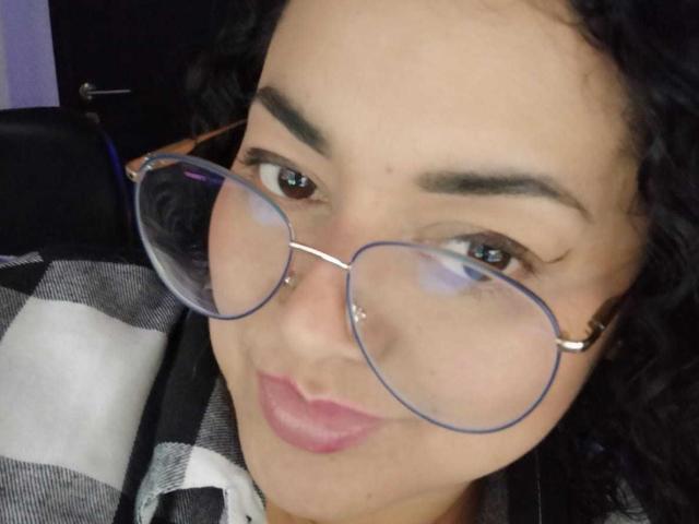 SophiLarusso with curly black hair, brown eyes, and pink lipstick, wearing blue glasses, has a nose piercing, and is smiling with a plump ass in a checkered shirt.