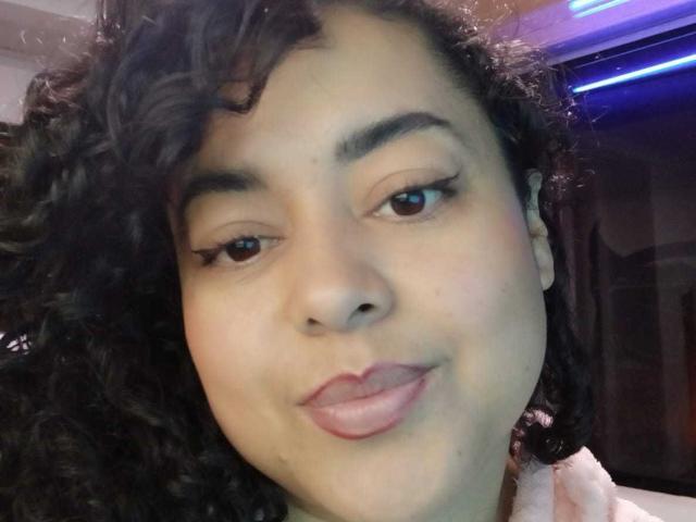 SophiLarusso with curly black hair, light brown skin, and subtle makeup, giving a slightly smirking, pouty look. She has small, dark brown eyes and pink lips.