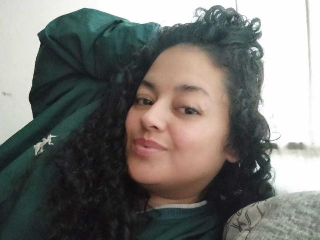 SophiLarusso with curly black hair, green shirt, and relaxed smile, hand behind head, showing soft brown skin and subtle makeup, slightly puffy lips.