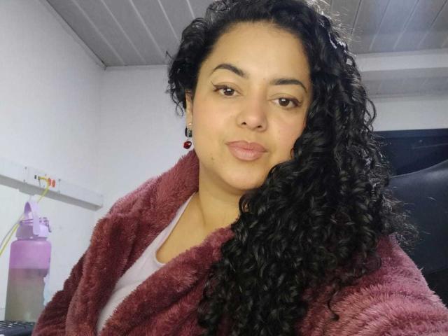SophiLarusso with curly black hair, brown eyes, and pink lips, wearing a red fuzzy robe over a white shirt, looking at the camera with a slight smile.