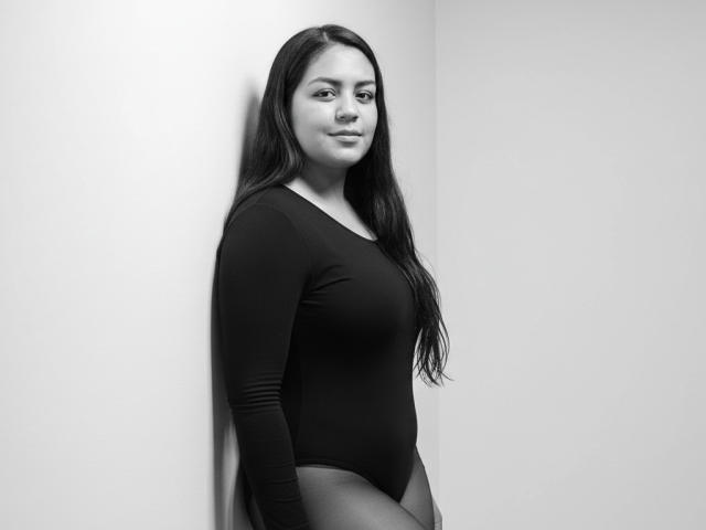 AthenaTheas, with long dark hair, wearing a black long-sleeve leotard, standing against a wall, smiling subtly, showing her tits and ass, in a black-and-white photo.