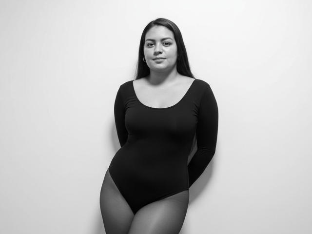 AthenaTheas with long dark hair, wearing a black long-sleeve bodysuit, standing against a white wall, hands behind her back, showcasing her tits and ass. Black-and-white photo.