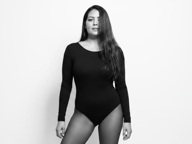 AthenaTheas with long dark hair, wearing a black long-sleeve leotard, standing, showing her ass, with a confident, slightly serious expression, in a black-and-white photo.