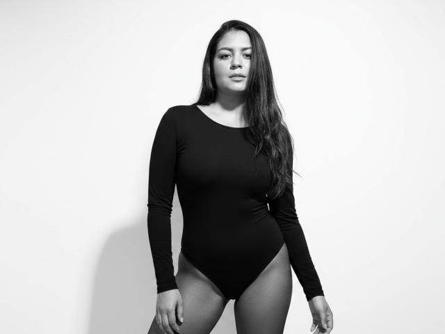 AthenaTheas with long dark hair, wearing a black long-sleeve bodysuit, showing her tits and ass, standing against a white wall, in a black-and-white photo.