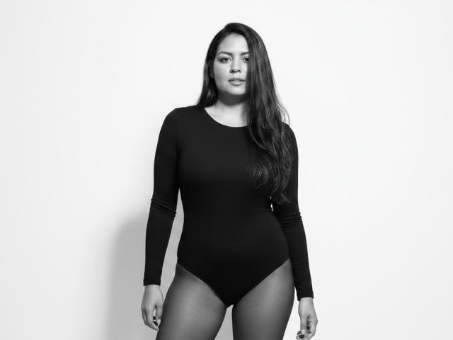 AthenaTheas with long dark hair, black long-sleeve leotard, and high-cut bottom, standing, showing her ass and small tits, looking at the camera in a black-and-white photo.